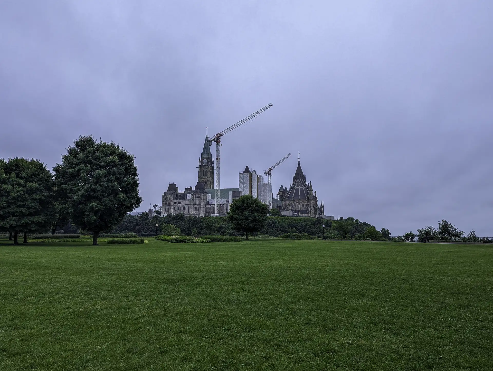 A grassy field with trees in the foreground leads to a large, historic building with towers and a crane in the background, under a cloudy sky. This scenic view captures the essence of Ottawa, the vibrant capital of Canada.